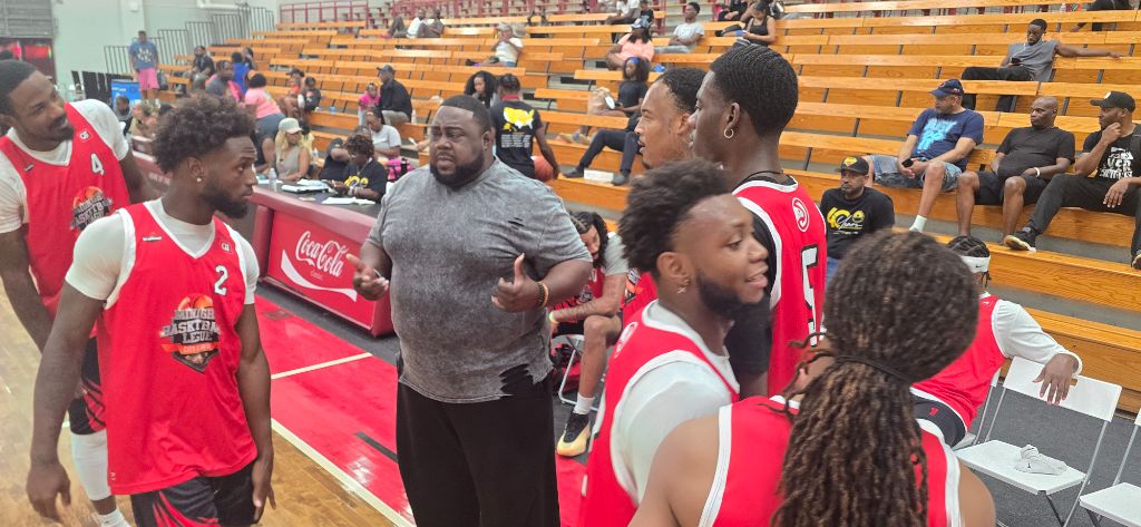 Coach in grey shirt addresses players in red Midnight Basketball League jerseys on the sideline at Forbes Arena — Coca-Cola scorer's table visible