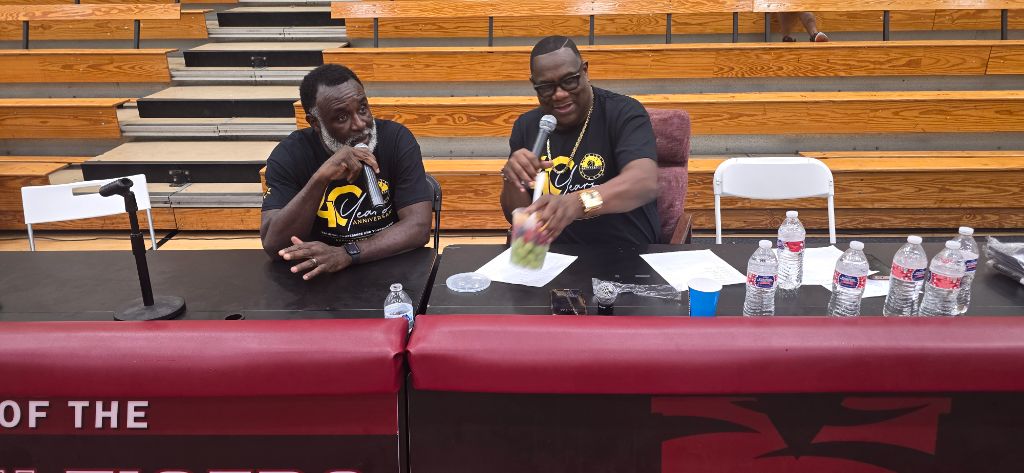 Two AMB leaders in 40th anniversary shirts at the panel table inside Forbes Arena — microphone, water bottles, and empty bleachers behind them