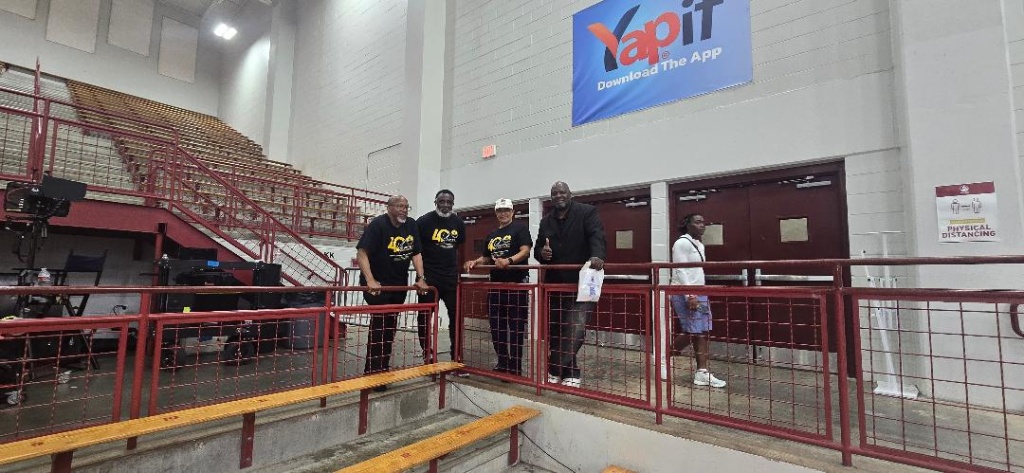 Five AMB leaders lean on the red railing inside Forbes Arena with wooden bleachers and a YapIt banner visible above